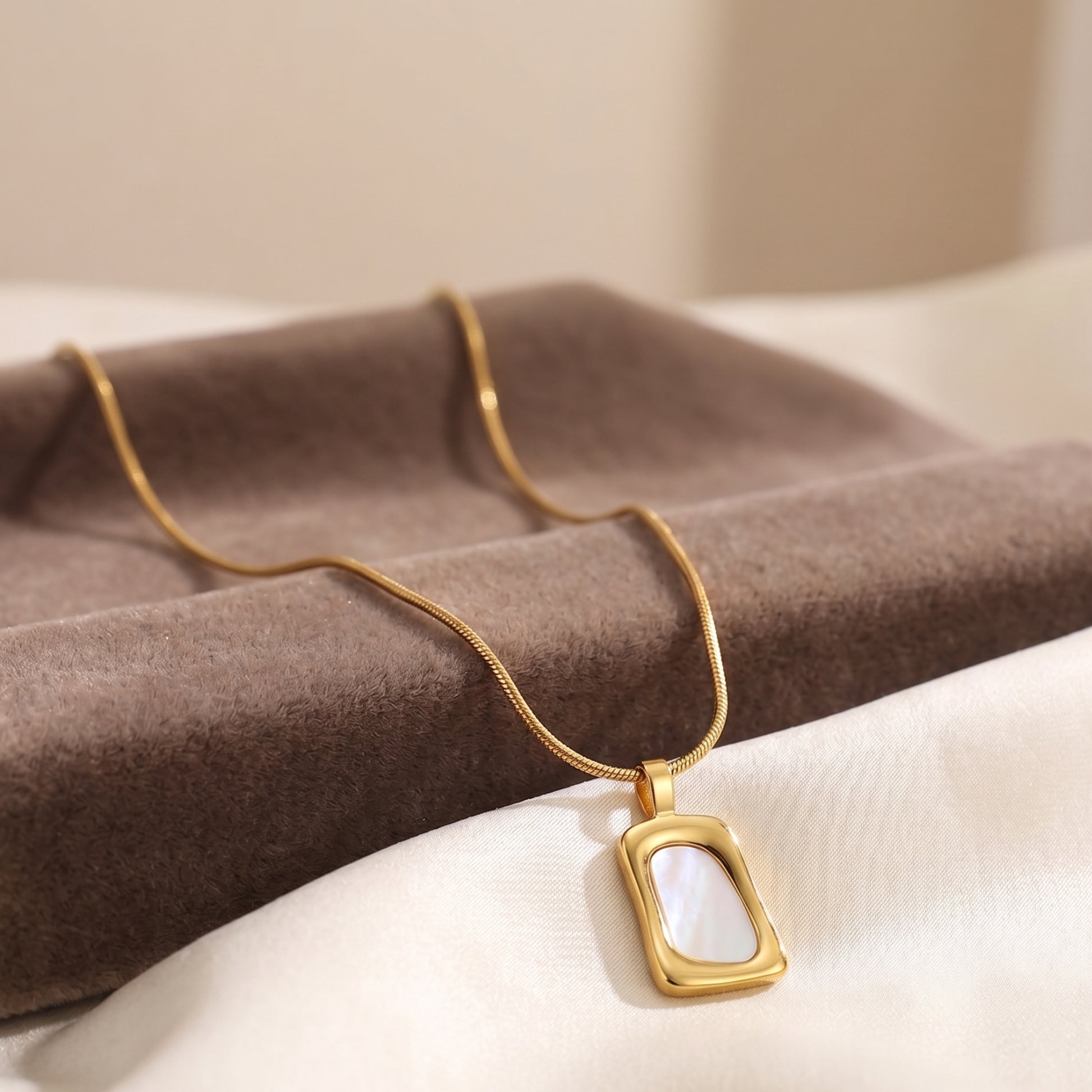 Close-up of a gold geometric pendant necklace with white mother of pearl inlay on a beige jewelry display box.