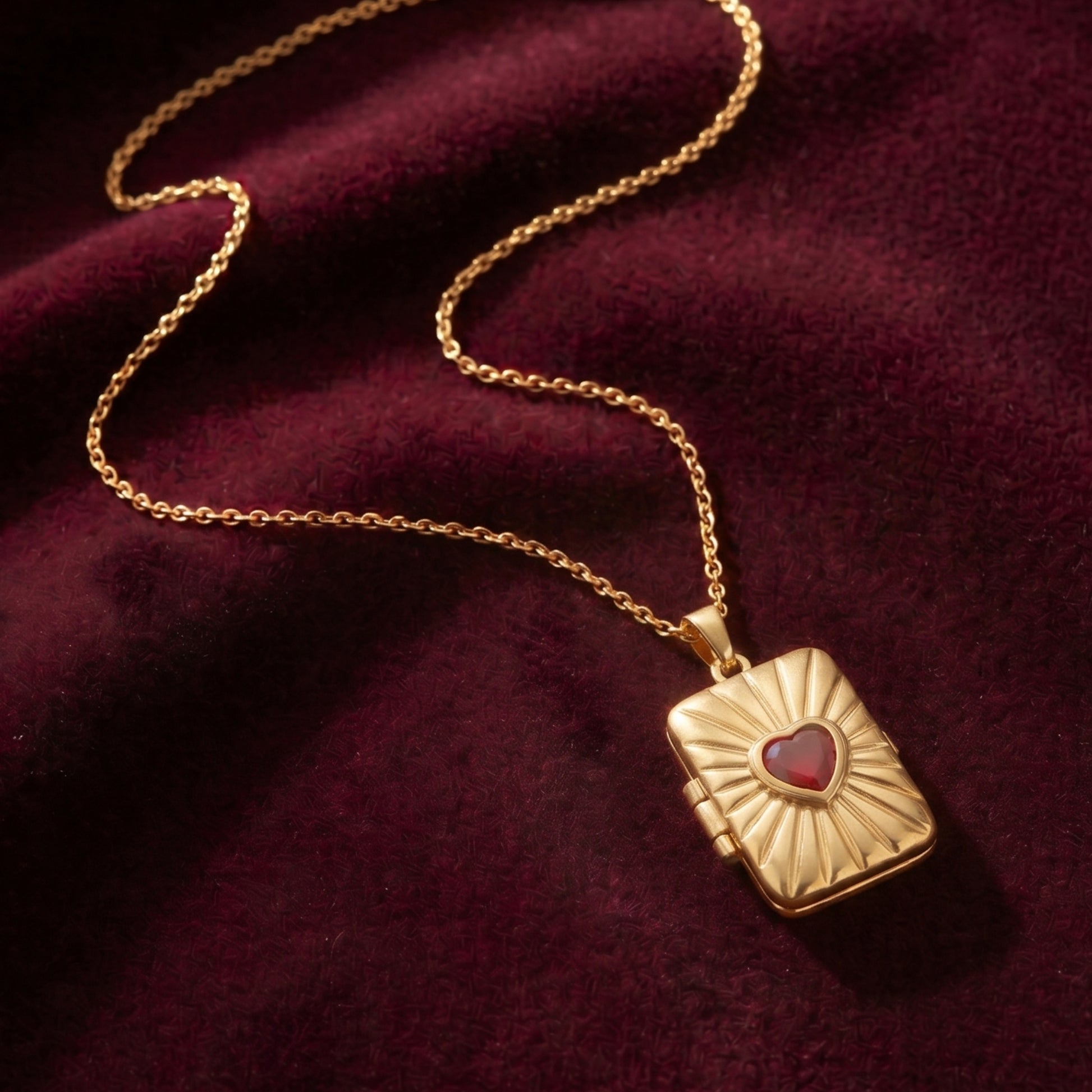 Close-up photograph of an gold-plated rectangular locket pendant with a radiant sunburst pattern and central heart-shaped red gemstone, resting on a deep burgundy velvet fabric with a gold chain.