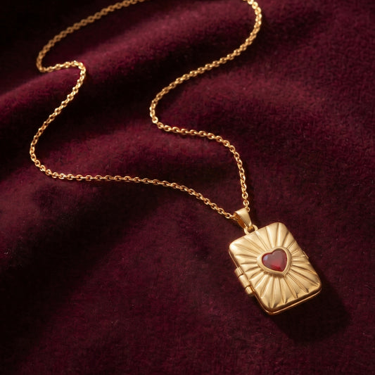 Close-up photograph of an gold-plated rectangular locket pendant with a radiant sunburst pattern and central heart-shaped red gemstone, resting on a deep burgundy velvet fabric with a gold chain.