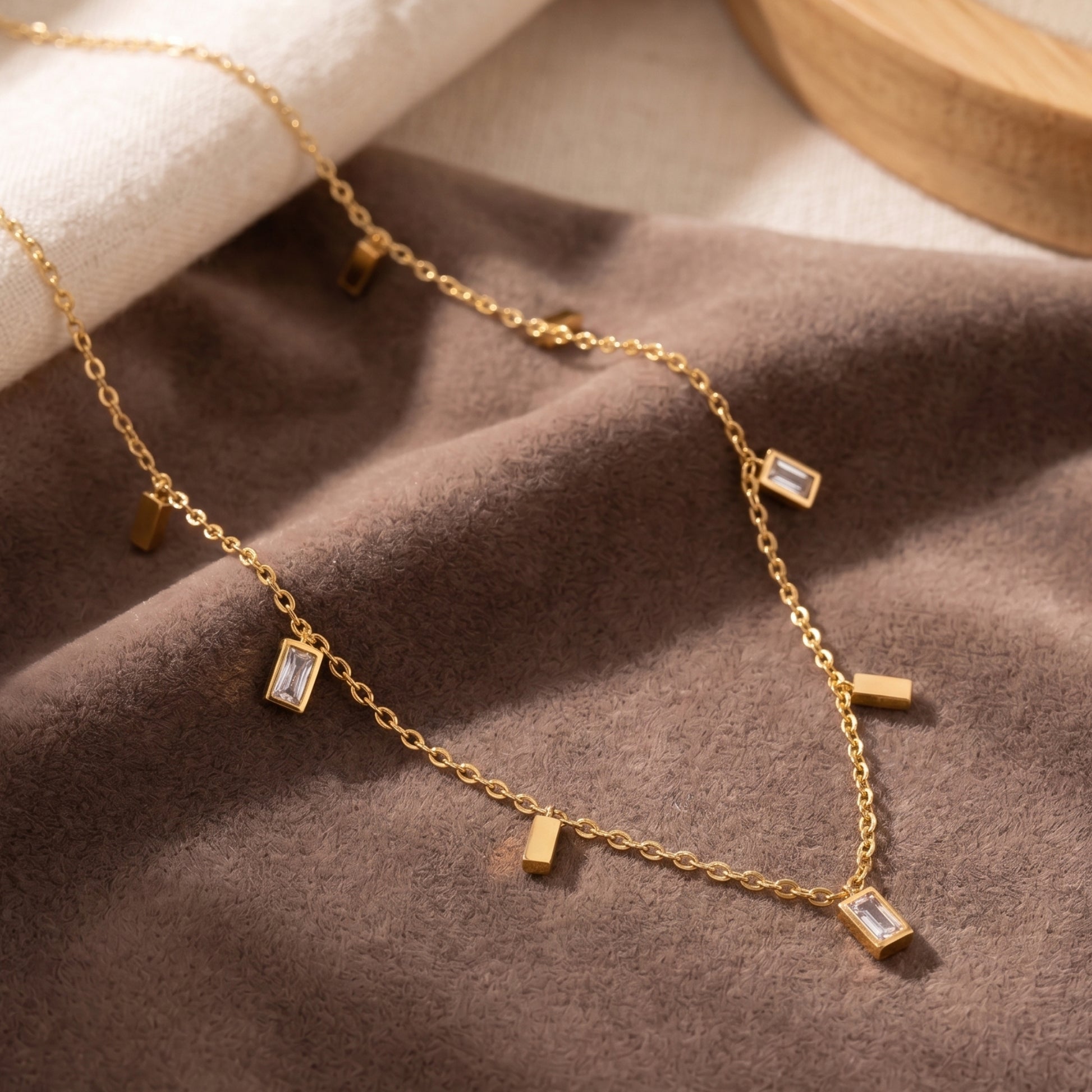 Close-up photograph of a dainty gold-plated station necklace on a beige jewelry display box, featuring alternating rectangular cubic zirconia baguette and polished gold bar dangles.