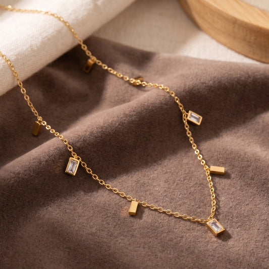 Close-up photograph of a dainty gold-plated station necklace on a beige jewelry display box, featuring alternating rectangular cubic zirconia baguette and polished gold bar dangles.