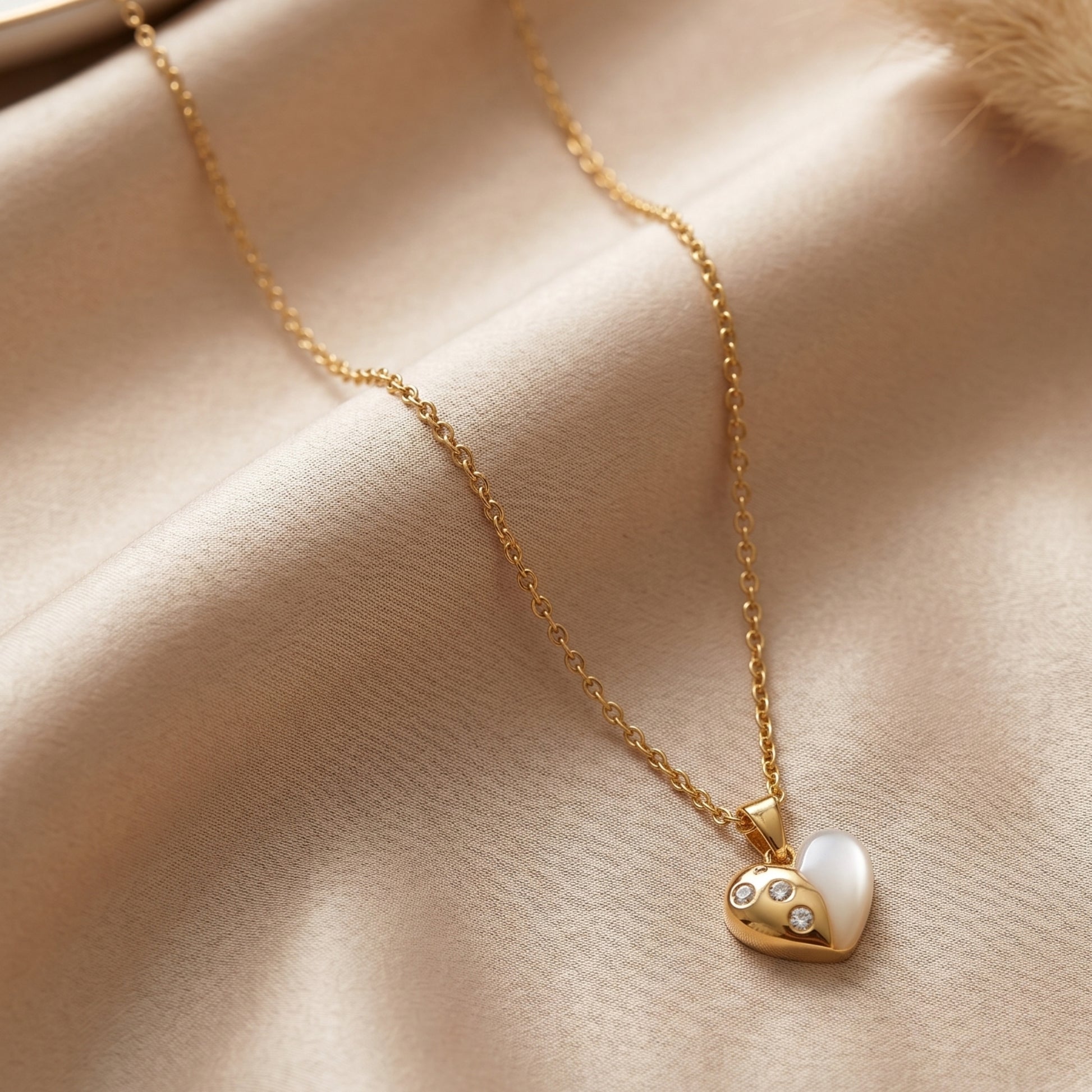 Close-up of a dainty gold heart pendant necklace with a white shell inlay and three small crystals on a soft beige silk background.