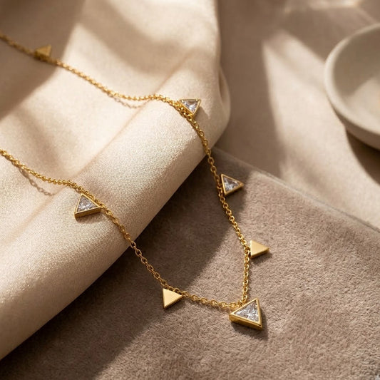 Dainty gold station necklace with alternating polished gold triangles and crystal-inlaid triangle charms, draped over silk and velvet fabric.