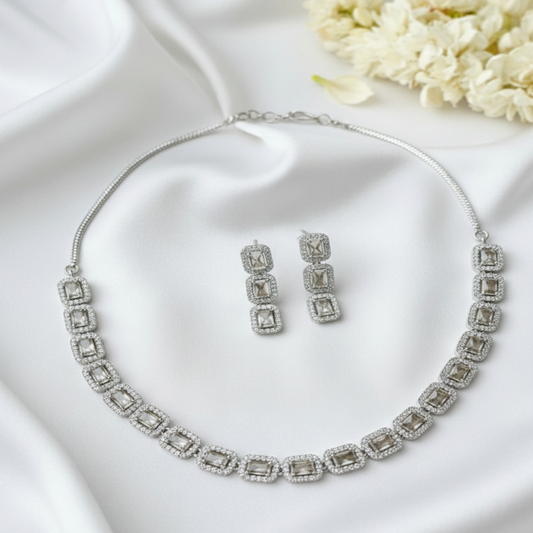 Silver necklace and earrings set on a white surface with flowers in the background