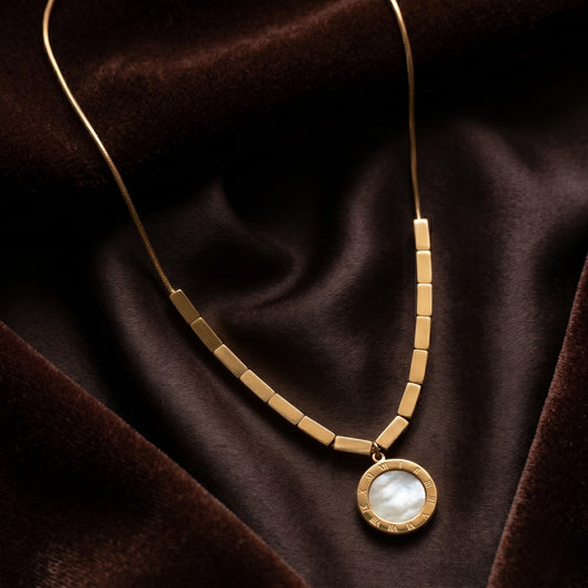 Close-up view of a gold-tone necklace with six rectangular tube beads and a circular pendant featuring a mother-of-pearl inlay and engraved Roman numerals, on a luxurious beige silk background.