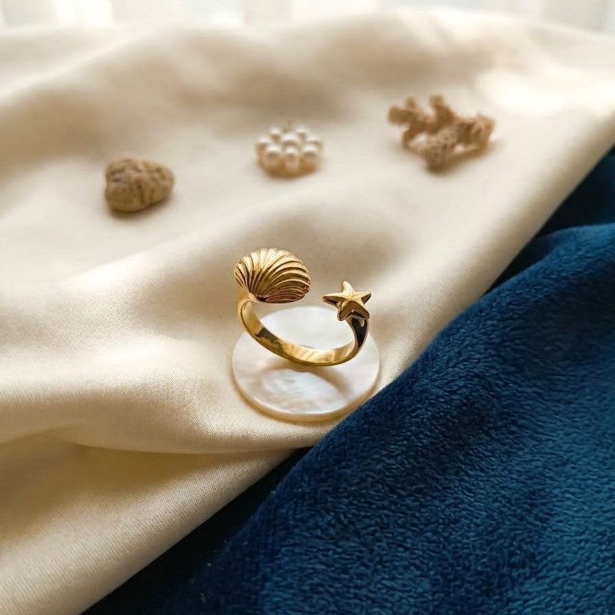 Adjustable gold-plated wrap ring featuring a seashell and starfish design, displayed on a mother-of-pearl base with cream and navy fabric.