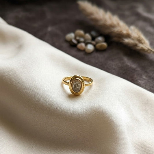 Dainty gold-plated ring with an oval-cut stone in a polished bezel setting, displayed on cream silk fabric with natural stone accents.